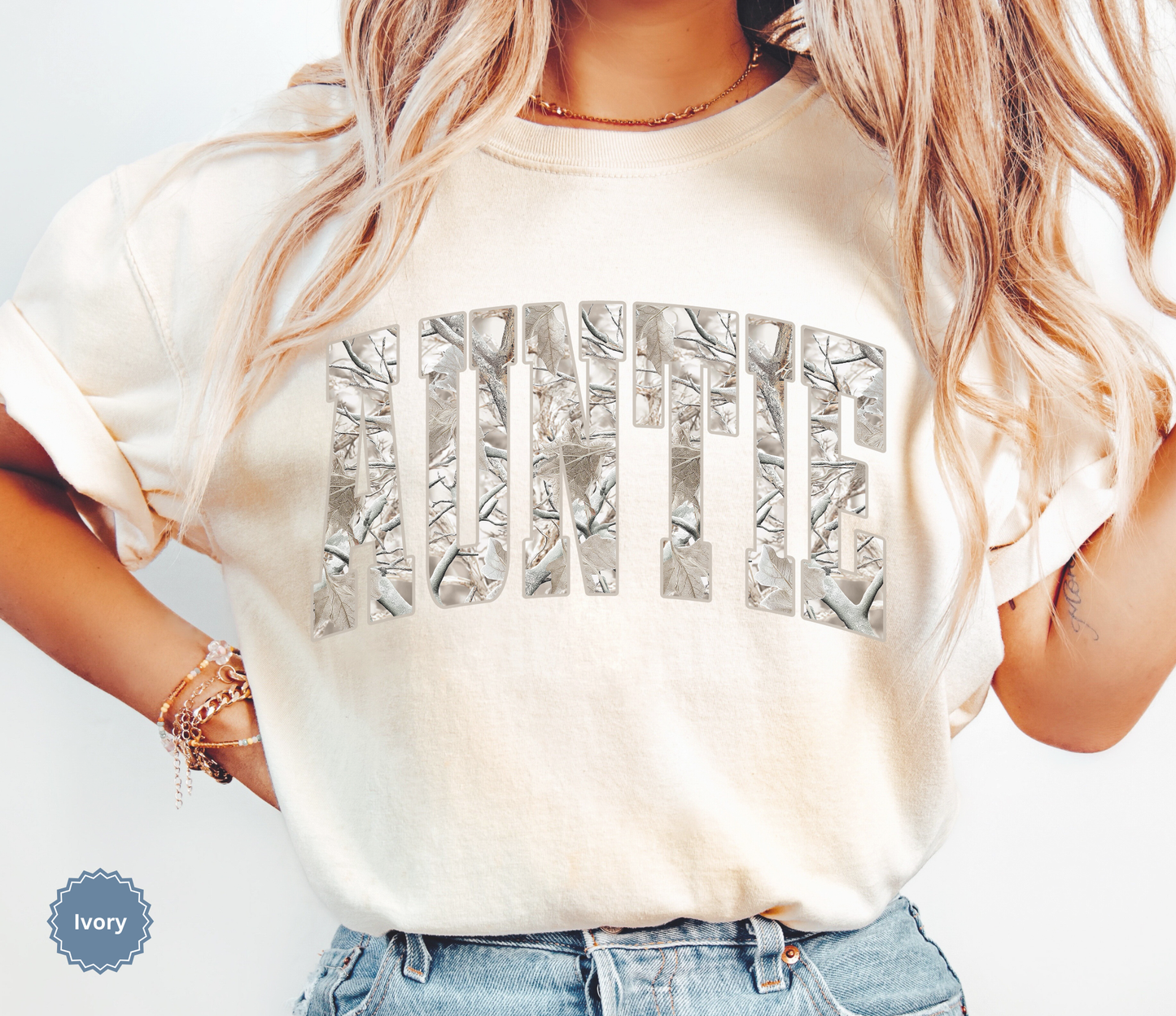 Woman wearing an ivory comfort colors t-shirt with 'Auntie' printed in a snow camo pattern.