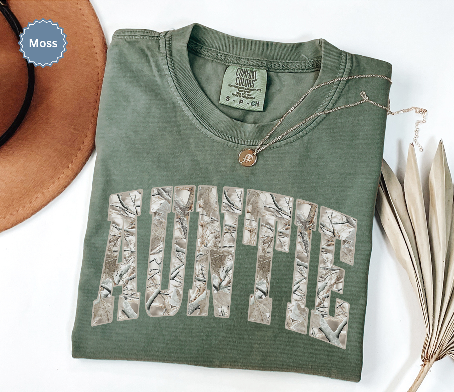 Folded comfort colors moss green t-shirt with 'Auntie' printed in a snow camo pattern.