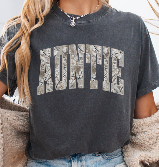 Woman wearing a comfort colors dark grey pepper t-shirt with 'Auntie' printed in a snow camo pattern.
