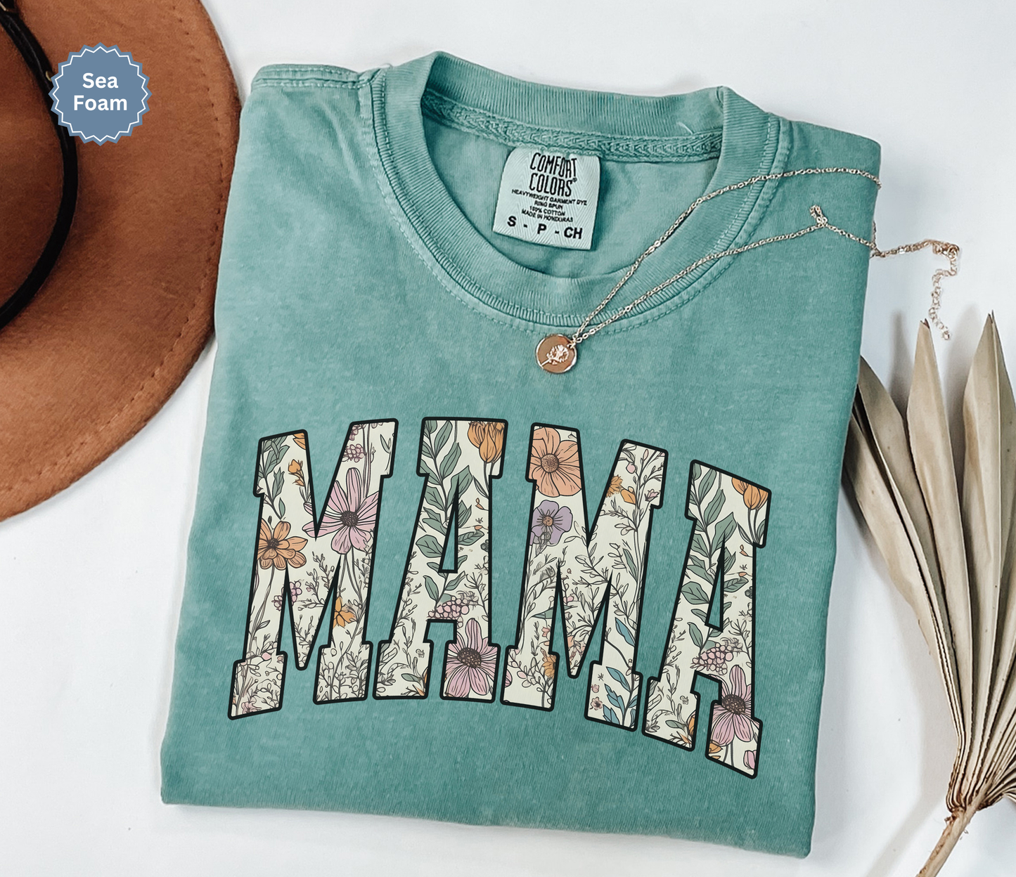 Folded seafoam green T-shirt with a varsity style "MAMA" floral print. 